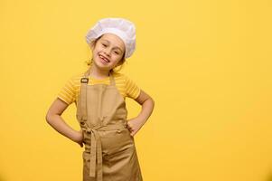 linda niño chica, pequeño cocinero confitero en delantal y blanco cocinero gorra, poniendo manos en cintura, sonriente mirando a cámara foto