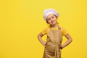 adorable pequeño niña en del chef delantal y sombrero, con manos en cintura, sonrisas mirando a cámara, aislado amarillo antecedentes foto
