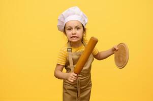 Adorable Caucasian little girl making faces, looking at camera, holding rolling pin and wooden board, isolated on yellow photo