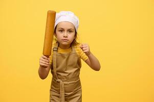 Serious angry little child girl in chef's uniform, holding a wooden rolling pin, clenching fists, looking at camera photo