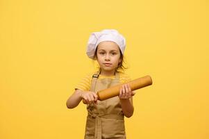 retrato de un bebé niña en del chef uniforme, participación un de madera laminación alfiler, mirando a cámara, aislado amarillo antecedentes foto