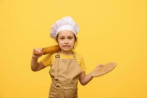 Beautiful Caucasian little girl making faces, looking at camera, holding rolling pin and wooden board, isolated yellow photo