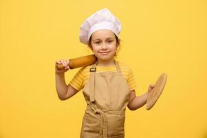 Adorable girl in chef hat and apron, smiles, looking at camera, holding rolling pin and wooden board, isolated on yellow photo