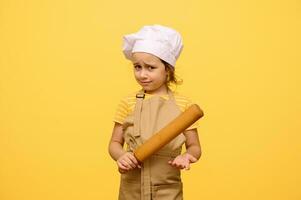 Studio portrait of amazed little girl, chef pastry, in white cap and beige apron, hoding rolling pin, isolated on yellow photo
