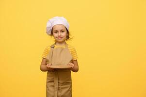 Charming adorable kid girl 6 years old, little chef pastry, smiles cutely and holds out at camera a wooden board photo