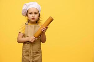 adorable pequeño niño niña en del chef sombrero y delantal, de pie con un laminación alfiler, aislado terminado amarillo estudio antecedentes foto