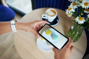 Female hands taking a photo of plate with macarons on a wooden table in cafeteria. Coffee break time. Mobile phone in live view mode