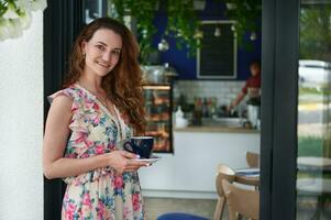 Young smiling woman standing with a cup of coffee in her hands at the entrance of her cafeteria photo