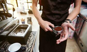 Partial view of unrecognizable barista in brown apron holding coffee press and portafilter with ground coffee photo