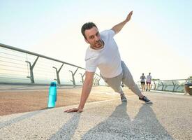 Middle aged Caucasian sportsman athlete doing push-ups, exercising on the glass city bridge at dawn. Bottle with water on the treadmill. Sport, active and healthy lifestyle concept photo