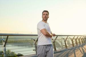 Middle aged European man in sportswear exercising outdoor on the city bridge at dawn on a warm sunny summer day. Sport, active and healthy lifestyle concept. Keep you body fit photo