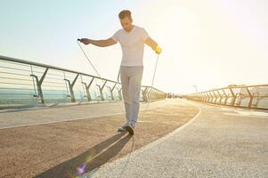 Full length portrait of a muscular build European middle aged man, fit athlete doing jumping exercises, cardio training with skipping rope on the urban city bridge early in the morning on a summer day photo