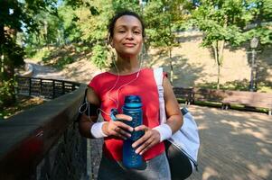 confidente atlético mujer en rojo apretado camiseta y terry pulseras sostiene botella con agua y sonrisas mirando a cámara foto