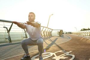 Middle-aged athlete, sportsman, muscular build European man is engaged in sports on a city bridge at dawn, performs squats, kneads muscles of the body. The concept of a healthy active lifestyle photo