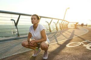 Portrait of charming middle aged Hispanic woman in white t-shirt and blue denim shorts squatting on treadmill and looking at camera while enjoying early morning walk at sunrise photo