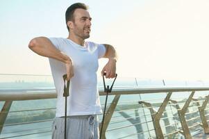 Handsome Caucasian middle aged man, athlete during outdoor workout with rubber resistance band. A middle-aged sportsman is training with an elastic expander on a glass city bridge at dawn photo