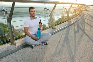 Handsome Caucasian muscular man, determined athlete, sportsman in headphones relaxing after workout outdoors at dawn, holding water bottle and sitting on glass city bridge in summer photo