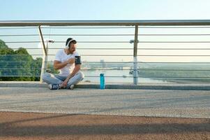 joven deportista, atleta en auriculares se sienta en un ciudad puente y cheques un móvil solicitud con supervisión corazón Velocidad y calorías quemado durante un rutina de ejercicio en su teléfono en un teléfono inteligente poseedor foto