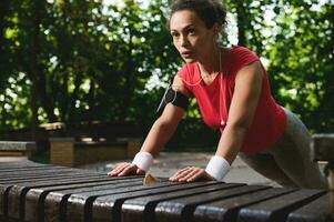 Determined Hispanic female athlete doing pushups from the wooden bench in the city park during functional cross training in the morning photo