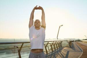 Handsome middle aged Caucasian man, athlete exercising outdoor, stretching body standing on a treadmill of a city bridge and warming up before morning jog at dawn photo