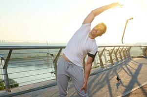 medio Envejecido muscular atleta estiramientos su brazo y hombro al aire libre y lo hace un calentamiento, disfrutando un Mañana rutina de ejercicio en un verano soleado día, en pie en un ciudad puente y admirativo el hermosa naturaleza foto