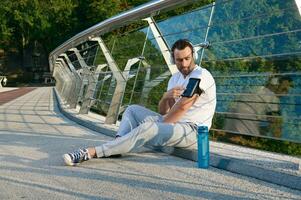 Attractive muscular man, athlete in headphones sits on a city bridge and checks a mobile application with monitoring heart rate and calories burned during a workout on his phone in a smartphone holder photo