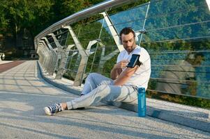 atractivo muscular hombre, atleta en auriculares se sienta en un ciudad puente y cheques un móvil solicitud con supervisión corazón Velocidad y calorías quemado durante un rutina de ejercicio en su teléfono en un teléfono inteligente poseedor foto