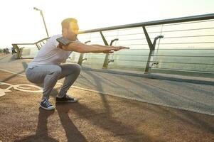 Middle aged Caucasian athlete doing deep squats on a city bridge on a sunny summer day. Concept of sport, movement, energy and dynamic, healthy lifestyle. photo