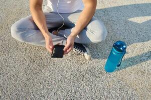 Top view of a sportsman wearing earphones sitting on the city bridge and checking his phone while relaxing after morning cardio workout and using a fitness tracking mobile application photo