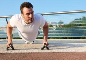 Close-up of a determined handsome middle aged man, athlete exercising outdoor, pumping muscles while performing push-ups training on the urban bridge on a beautiful summer day photo