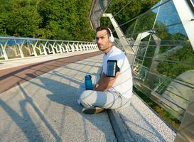 Confident relaxed handsome European man, athlete in sportswear and wireless headphones, listening to the music while relaxing on the urban glass bridge after morning workout on a warm summer sunny day photo