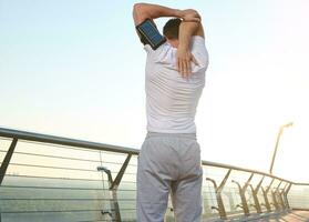 Athlete stretching his arm and shoulder outdoors, doing a warm-up, enjoying an early morning workout at dawn, standing on a city bridge and admiring the beautiful nature. Rear view photo