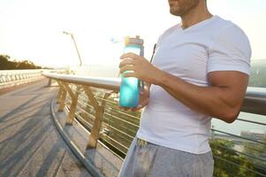 Cropped view of a muscular build European man, fit athlete with beautiful aesthetic body holding a bottle with water for rehydration and refreshment after heavy outdoor workout at dawn photo