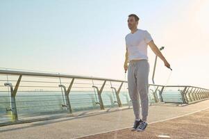 Full length portrait of a muscular build Caucasian man, fit athlete doing jumping exercises, cardio training with a skipping rope on the glass city bridge early in the morning on a summer day photo