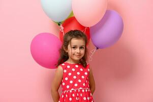 maravilloso bebé niña vistiendo verano polca puntos vestido, participación multicolor globos detrás su atrás, mirando a cámara. contento mujer y muchachas día concepto. cumpleaños, festivales eventos, celebraciones foto