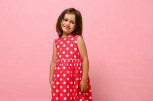 maravilloso europeo bebé chica, adorable niño vestido en rosado lunares vestido, linda sonrisas posando terminado rosado antecedentes con espacio para texto. confidente retrato de un hermosa caucásico niño niña foto