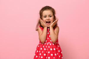 Close-up portrait of a gorgeous baby girl in a pink dress with polka dots poses at the camera, standing against a pink background and holding her chin with her hands. photo