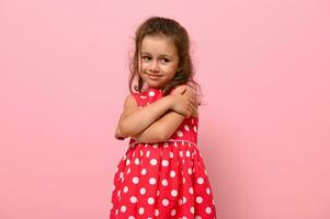 maravilloso tímidamente 4 4 años niño en Lunares vestir abrazos sí misma, dulce sonrisas posando en rosado pared antecedentes con espacio para texto. confidente retrato de hermosa bebé niña para publicidad foto