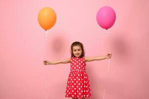 adorable maravilloso 4 4 años bebé niña en rosado vestir con blanco polca puntos, participación vistoso globos en su extendido manos, sonriente mirando a el cámara, aislado en rosado antecedentes con Copiar espacio foto