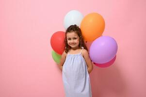 Advertising photography with cute baby girl in summer dress holding colorful balloons, posing against pink background, copy space photo