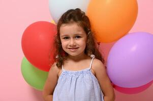 Isolated portrait, head shot, close-up of beautiful cheerful 4 years baby girl with multicolored balloons smiling looking at camera. Pink background copy space photo