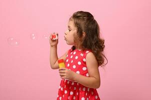 Side portrait of a cheerful beautiful baby girl blowing soap bubbles, isolated over pink background with copy space. Gorgeous child playing with soap bubbles. Summer children leisures concept photo
