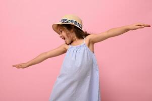 Gorgeous Caucasian ethnicity 4 years baby girl in summer blue dress and straw hat posing with outstretched arms looking down on a pink background with space for text photo