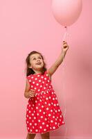 Adorable gorgeous 4 year pretty birthday girl in pink polka dot dress rejoices looking at pink balloon in her raised hand, isolated on pink background with copy space. True childhood emotions.Concepts photo