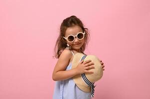 Beautiful shyly baby girl in sunglasses cute smiles posing over pink background with a straw summer hat in her hands. Summer concepts with copy space photo