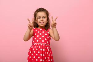 Joyful gorgeous 4 years baby girl in summer dress gesturing, doing victory sign with finger. Adorable child showing number two. photo