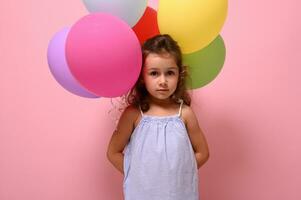 Confident portrait of beautiful little girl holding multicolored balloons behind back, looking at camera posing against pink background with copy space photo