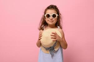 adorable 4 4 años niño vistiendo azul verano vestir y Gafas de sol, poses a cámara con Paja sombrero en su manos. aislado retrato de hermosa bebé niña en rosado antecedentes con Copiar espacio foto