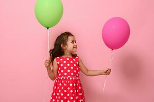 hermosa maravilloso 4 4 año bonito cumpleaños niña en rosado polca punto vestido, sostiene dos multicolor globos, se regocija mirando a un verde globo en su mano, aislado en rosado antecedentes con Copiar espacio. foto