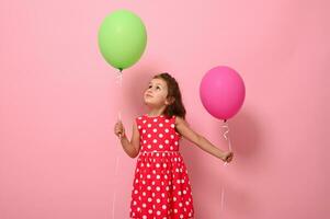 Adorable gorgeous 4 year pretty birthday girl in pink polka dot dress, holds two multicolored balloons, rejoices looking at a green balloon in her hand, isolated on pink background with copy space. photo